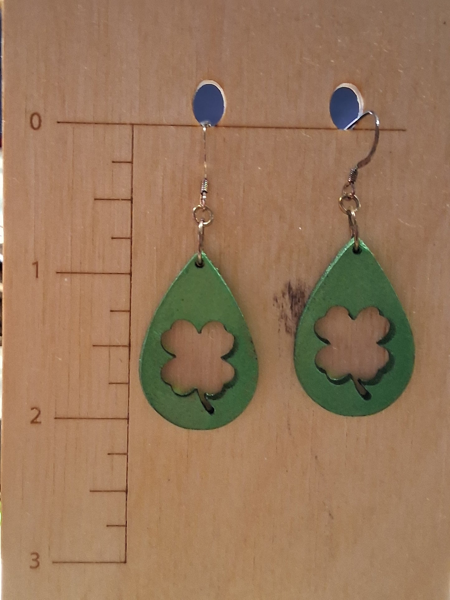 Green teardrop earrings with a clover design on a wooden surface with a ruler for scale.