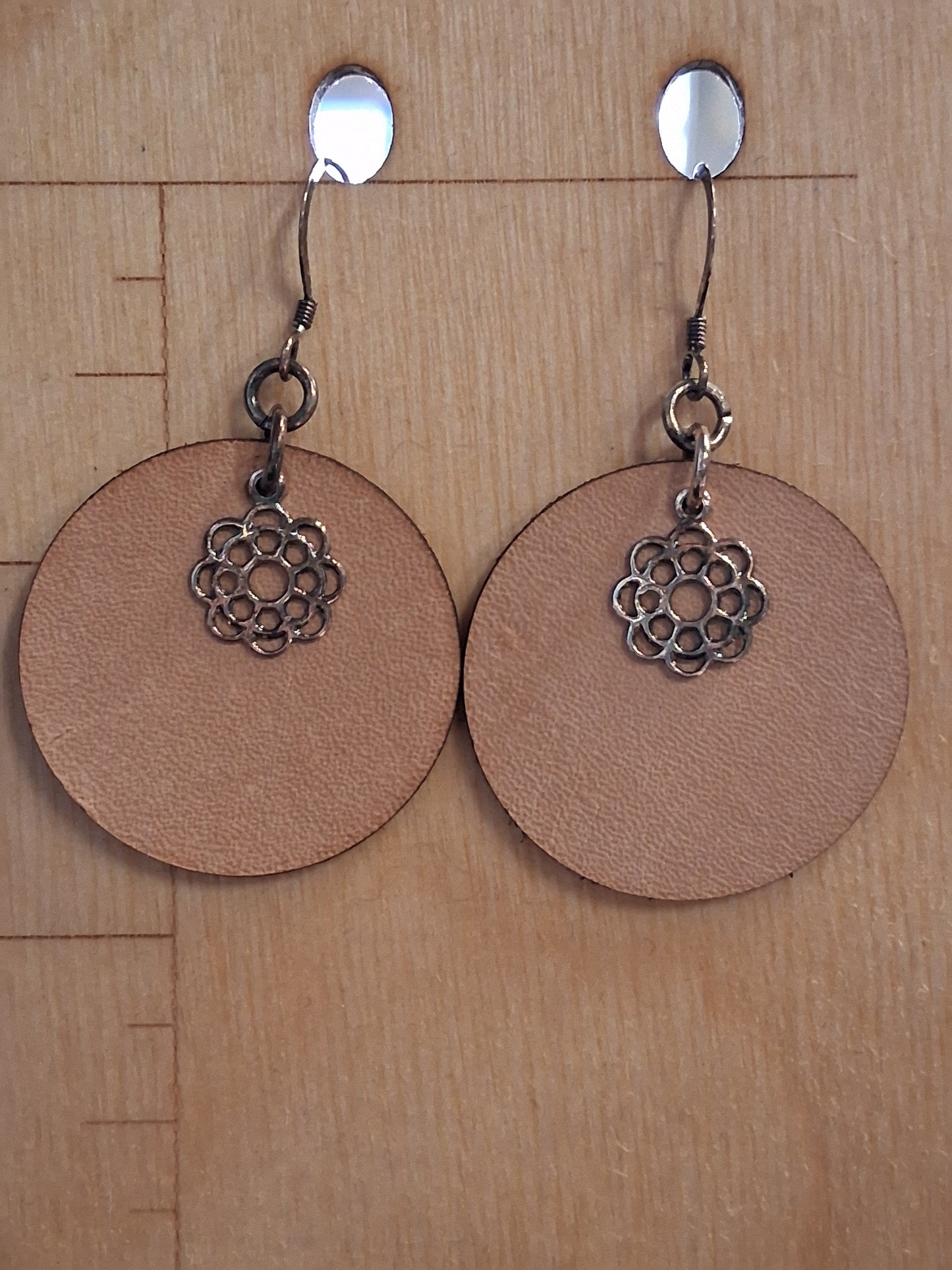 Pair of earrings with circular brown leather and floral charm on a wooden surface with measurement markings.
