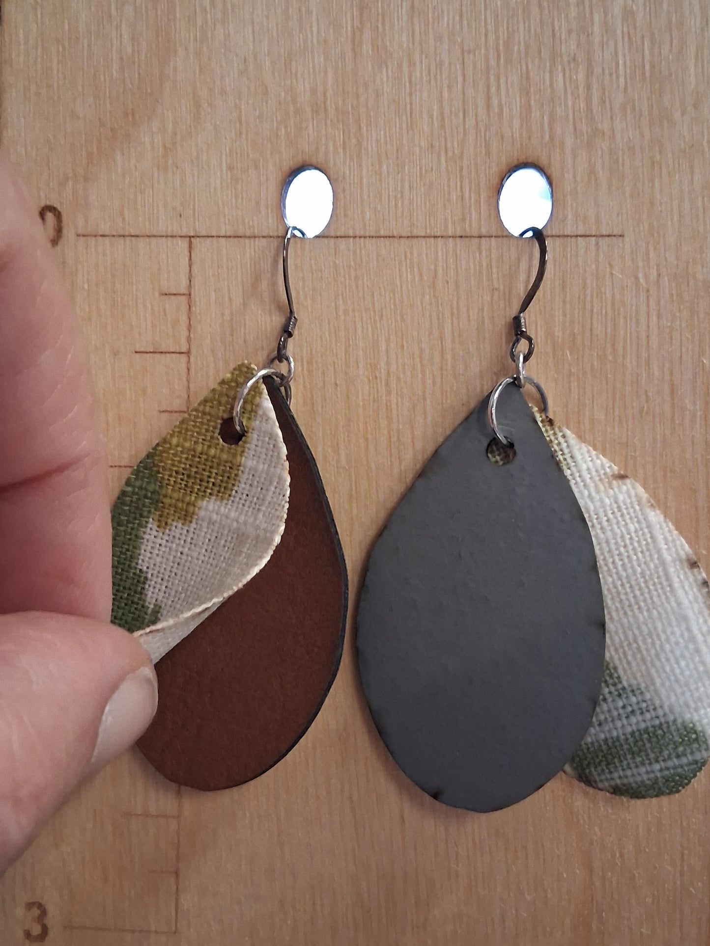 Pair of earrings with camouflage design fabric in front of a pleather back on a wooden surface with a ruler for scale.
