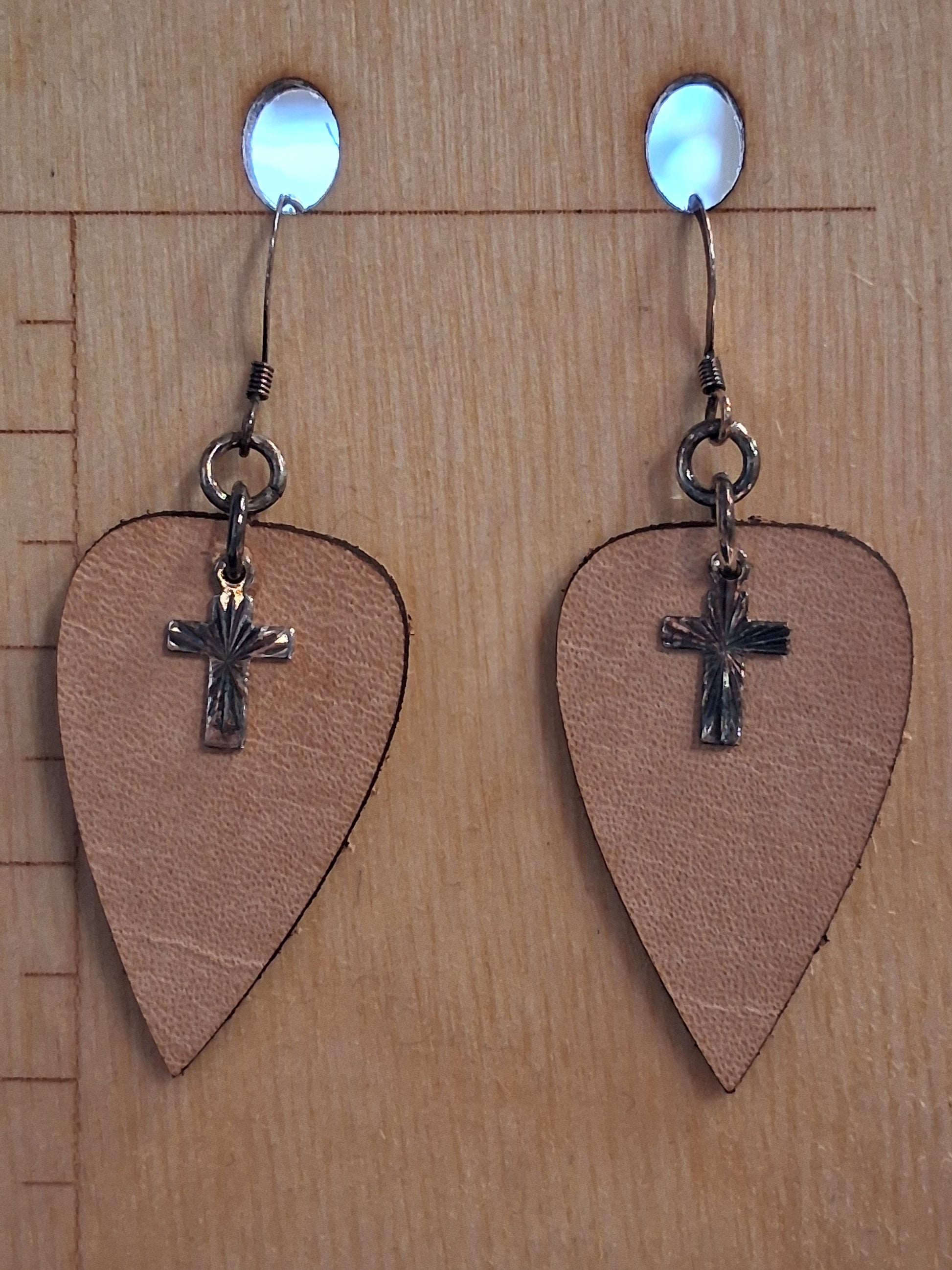 Pair of leather upside-down teardrop earrings with silver cross charms on a wooden surface 