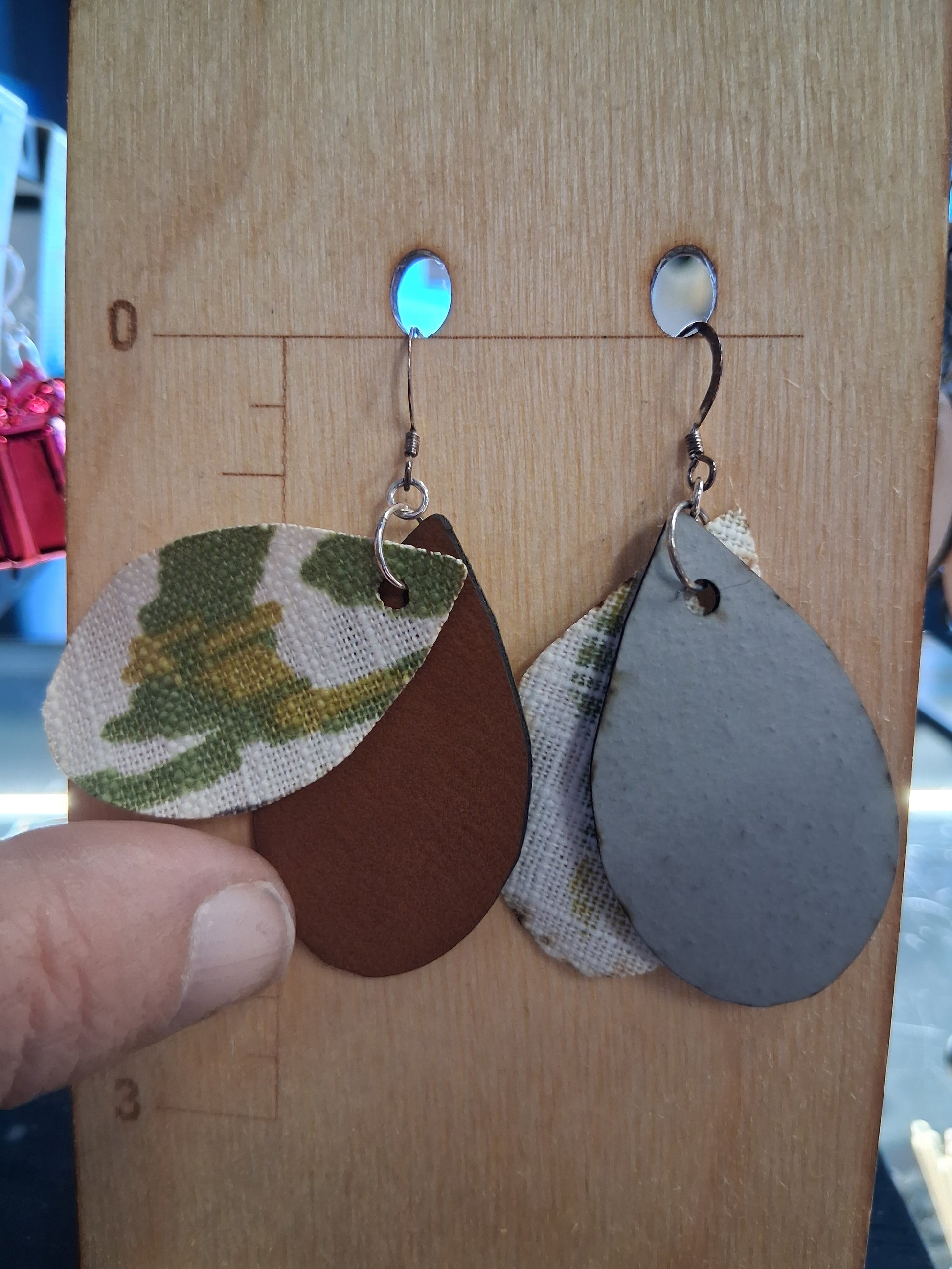Pair of earrings with camouflage design fabric in front of a pleather back on a wooden surface with a ruler for scale.
