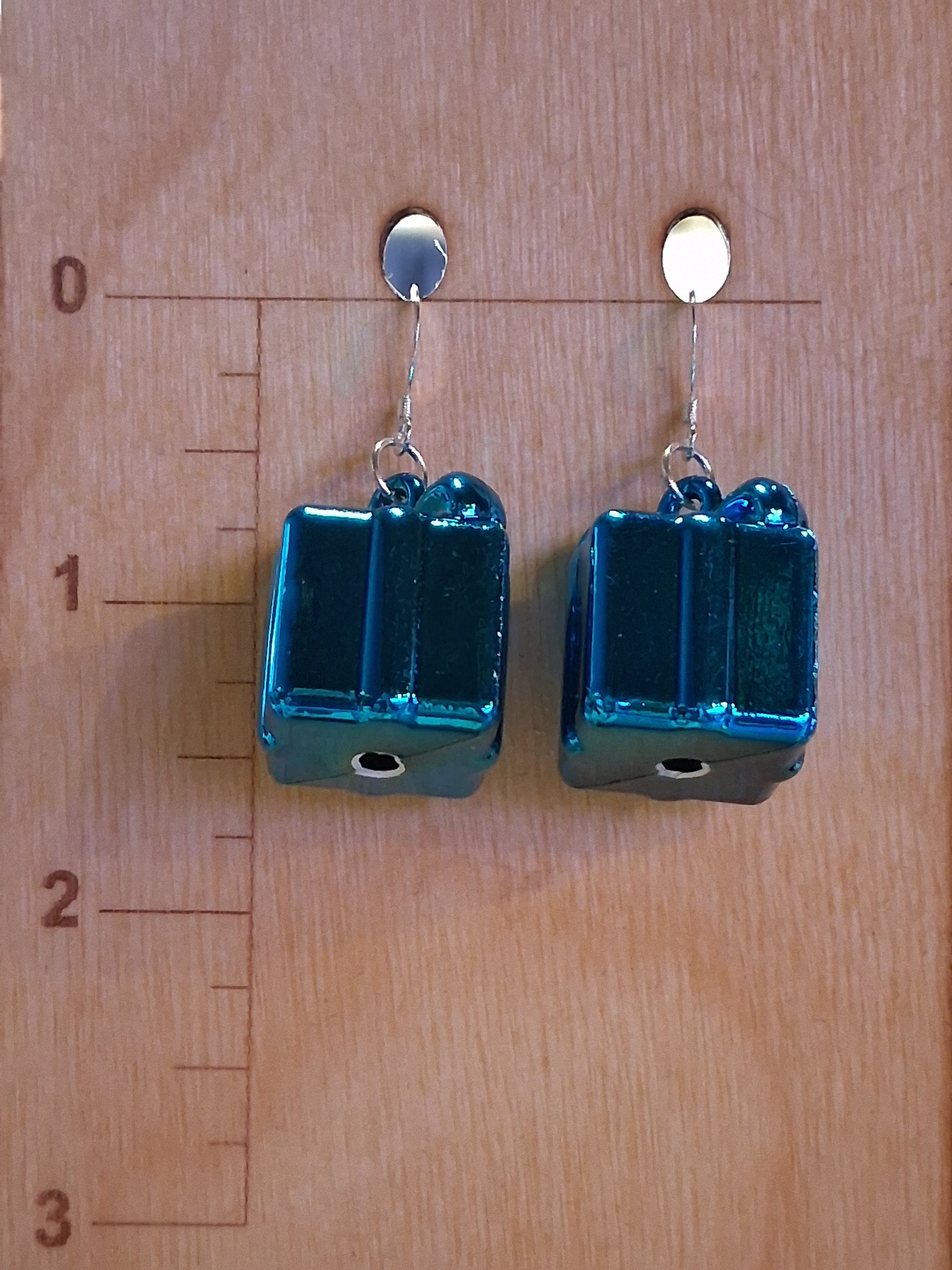 Close-up of metallic blue present-shaped dangle earrings displayed for scale on a wooden jewelry board. Blue rectangular present earrings on a wooden surface with a ruler for scale. Pair of sapphire blue Christmas gift charm earrings featuring reflective finish and silver-tone findings. 