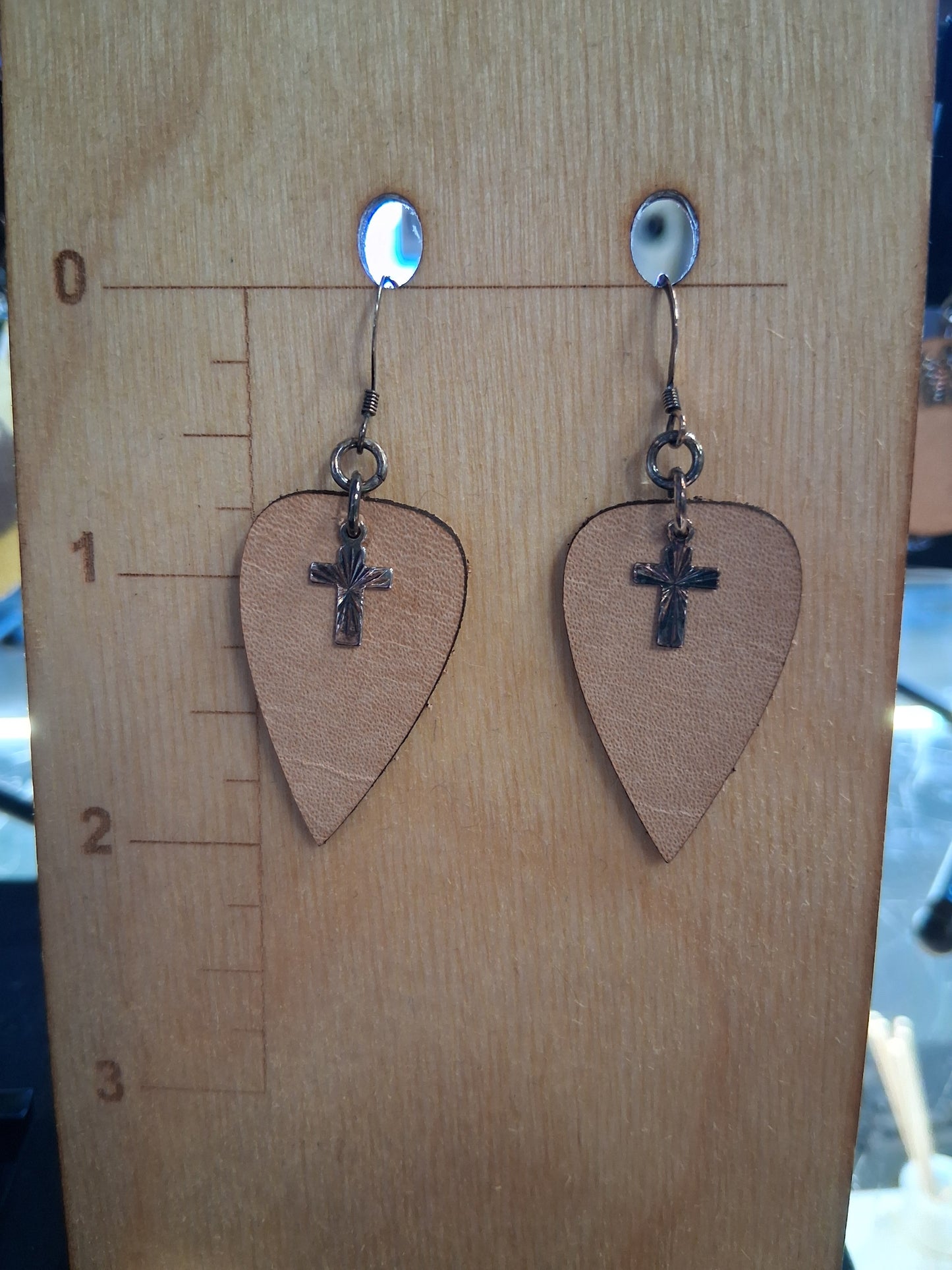 Pair of leather upside-down teardrop earrings with silver cross charms on a wooden surface with a ruler for scale.