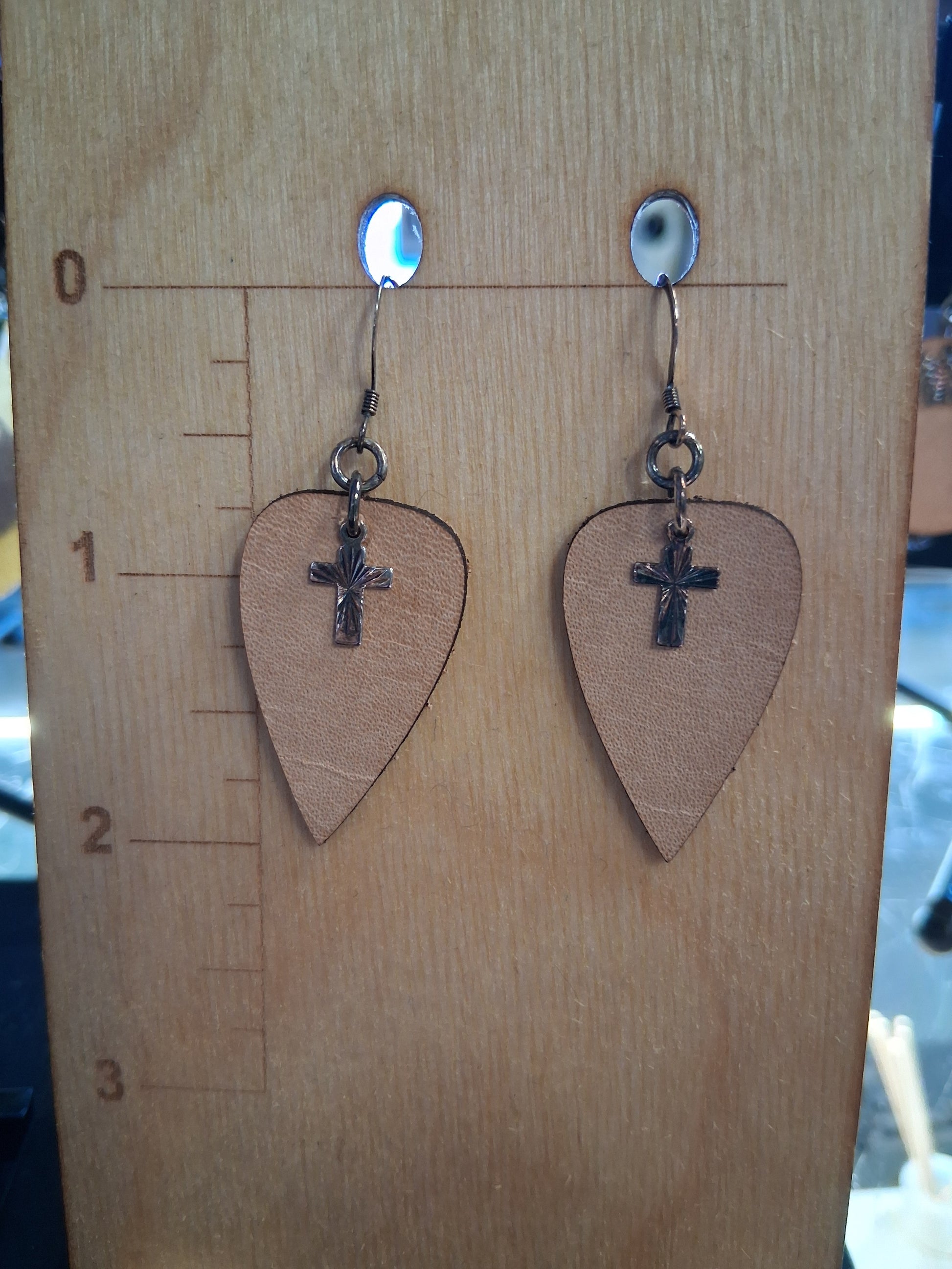 Pair of leather upside-down teardrop earrings with silver cross charms on a wooden surface with a ruler for scale.