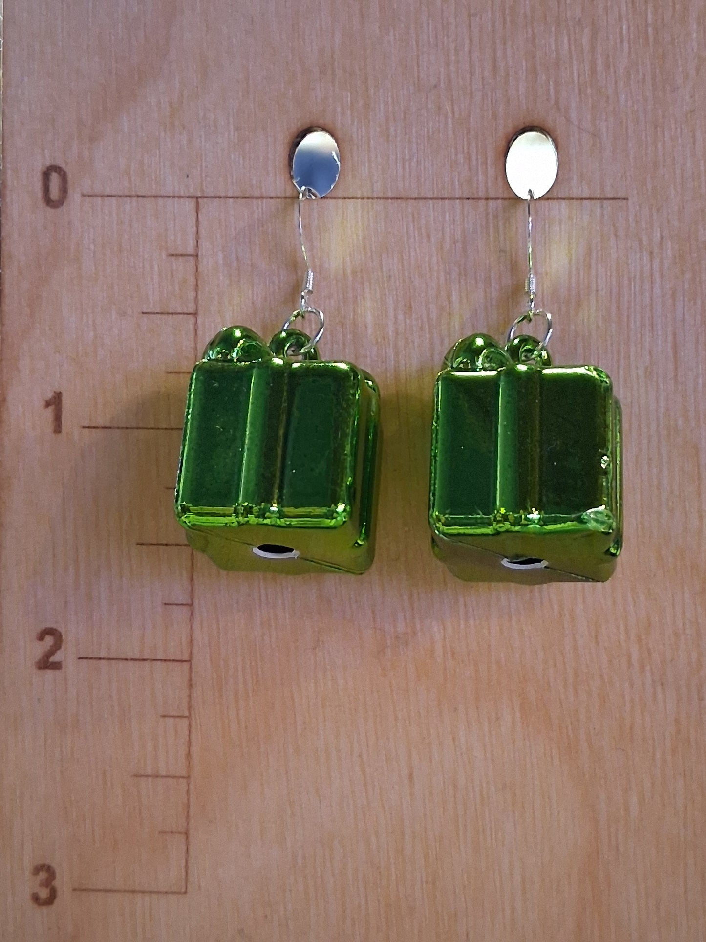 Close-up of green Christmas present charm earrings featuring metallic gift box charms and dark metal findings. Green rectangular present earrings on a wooden surface with a ruler for scale.