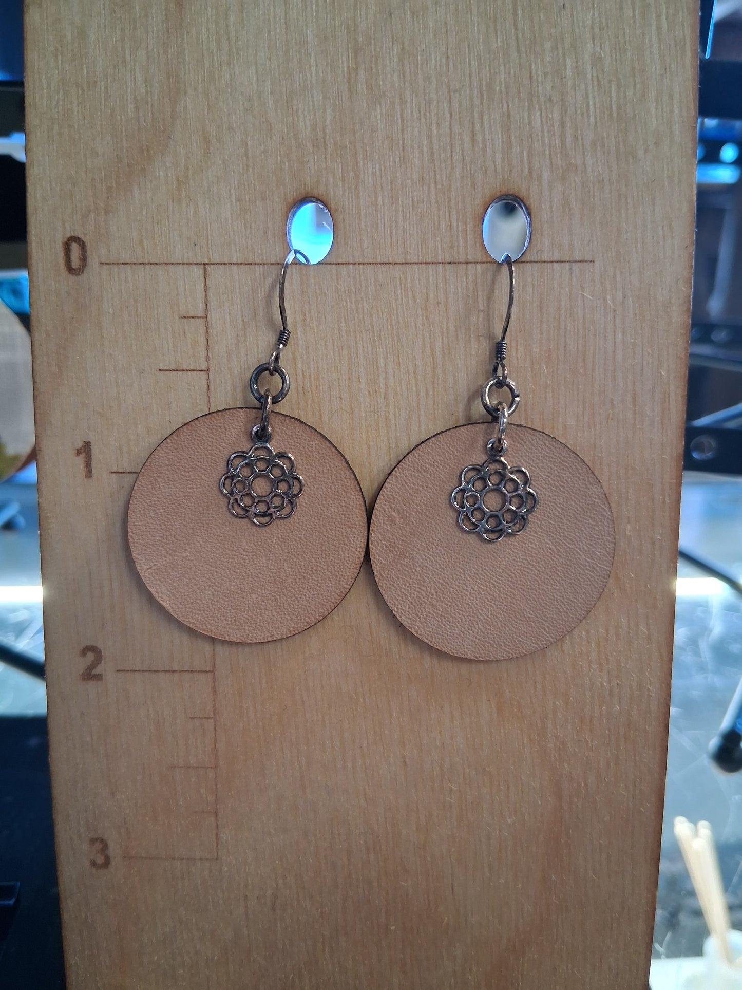 Pair of earrings with circular brown leather and floral charm on a wooden surface with measurement markings.