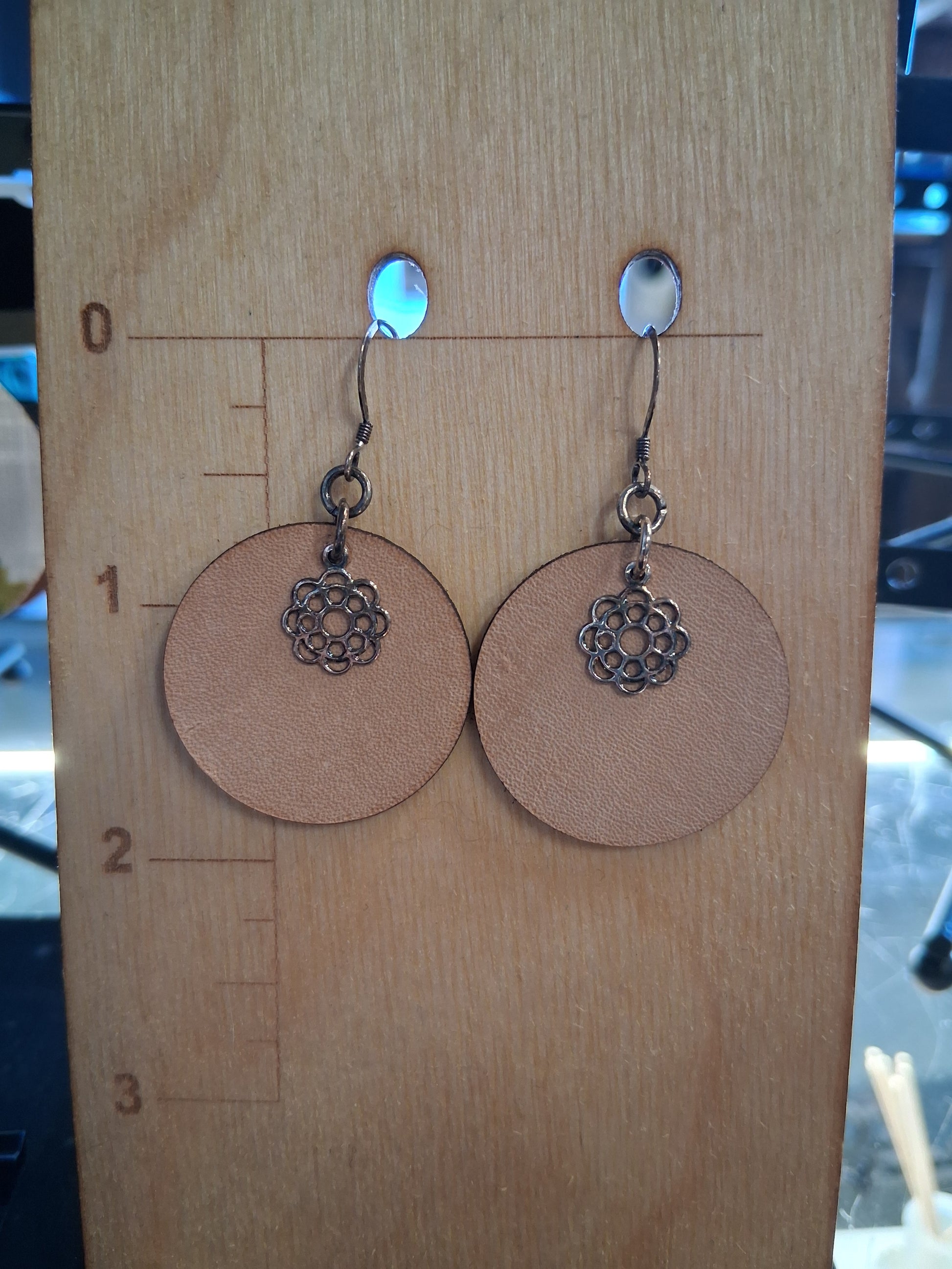 Pair of earrings with circular brown leather and floral charm on a wooden surface with measurement markings.