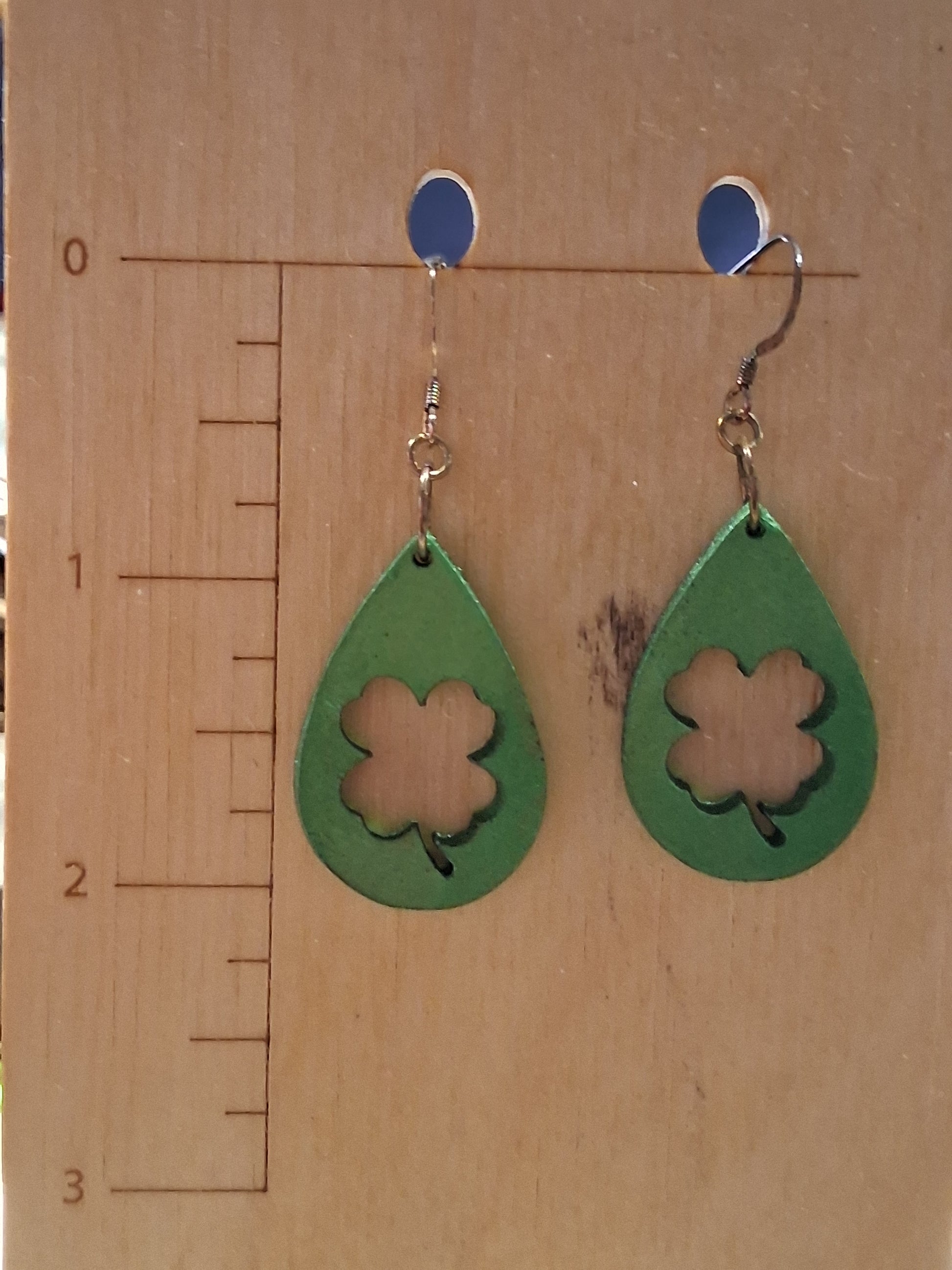 Green teardrop earrings with a clover design on a wooden surface with a ruler for scale.