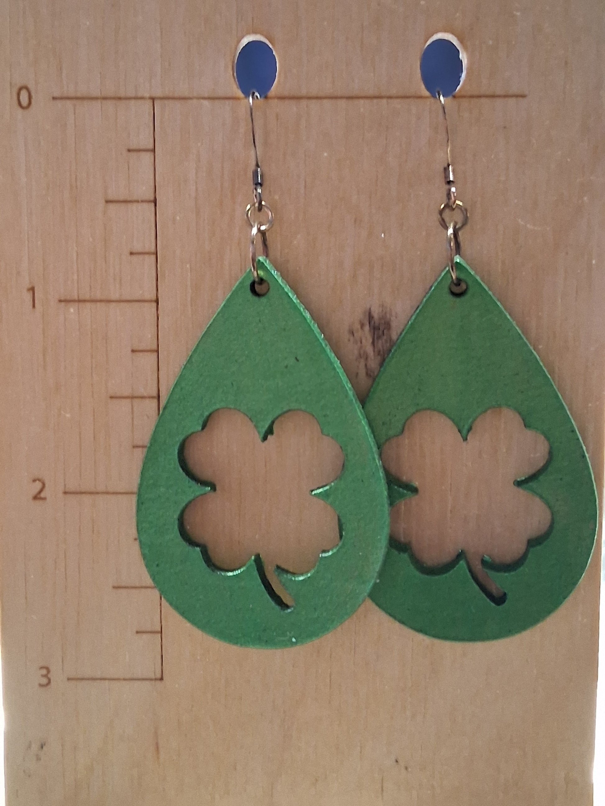 Green teardrop-shaped earrings with shamrock cutouts on a wooden surface with a ruler.