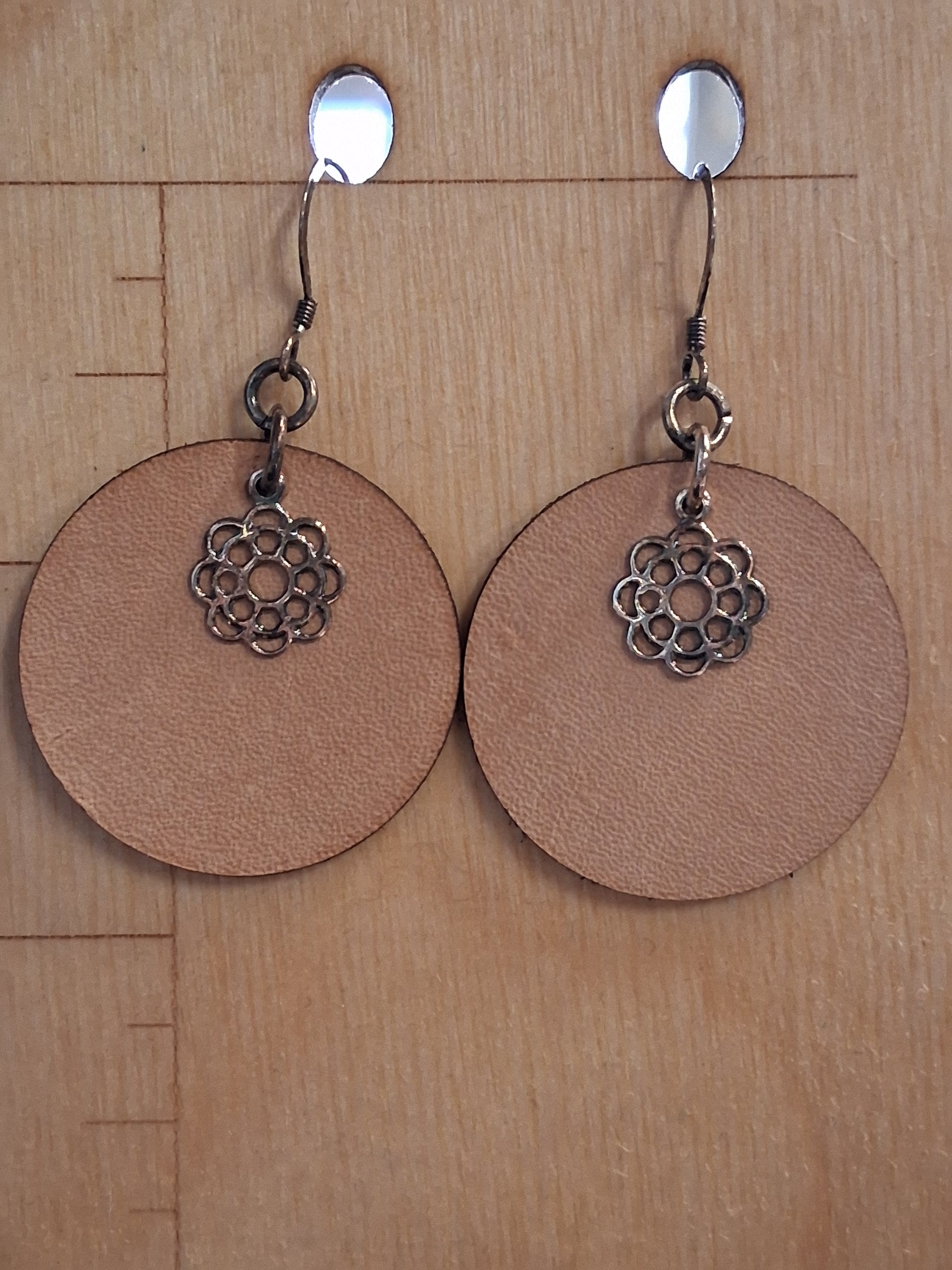 Pair of earrings with circular brown leather and floral charm on a wooden surface with measurement markings.