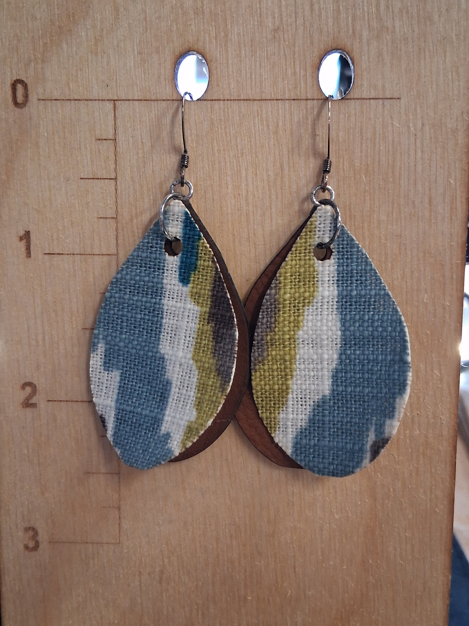 Pair of earrings with fabric in front of a pleather back on a wooden surface with a ruler for scale.