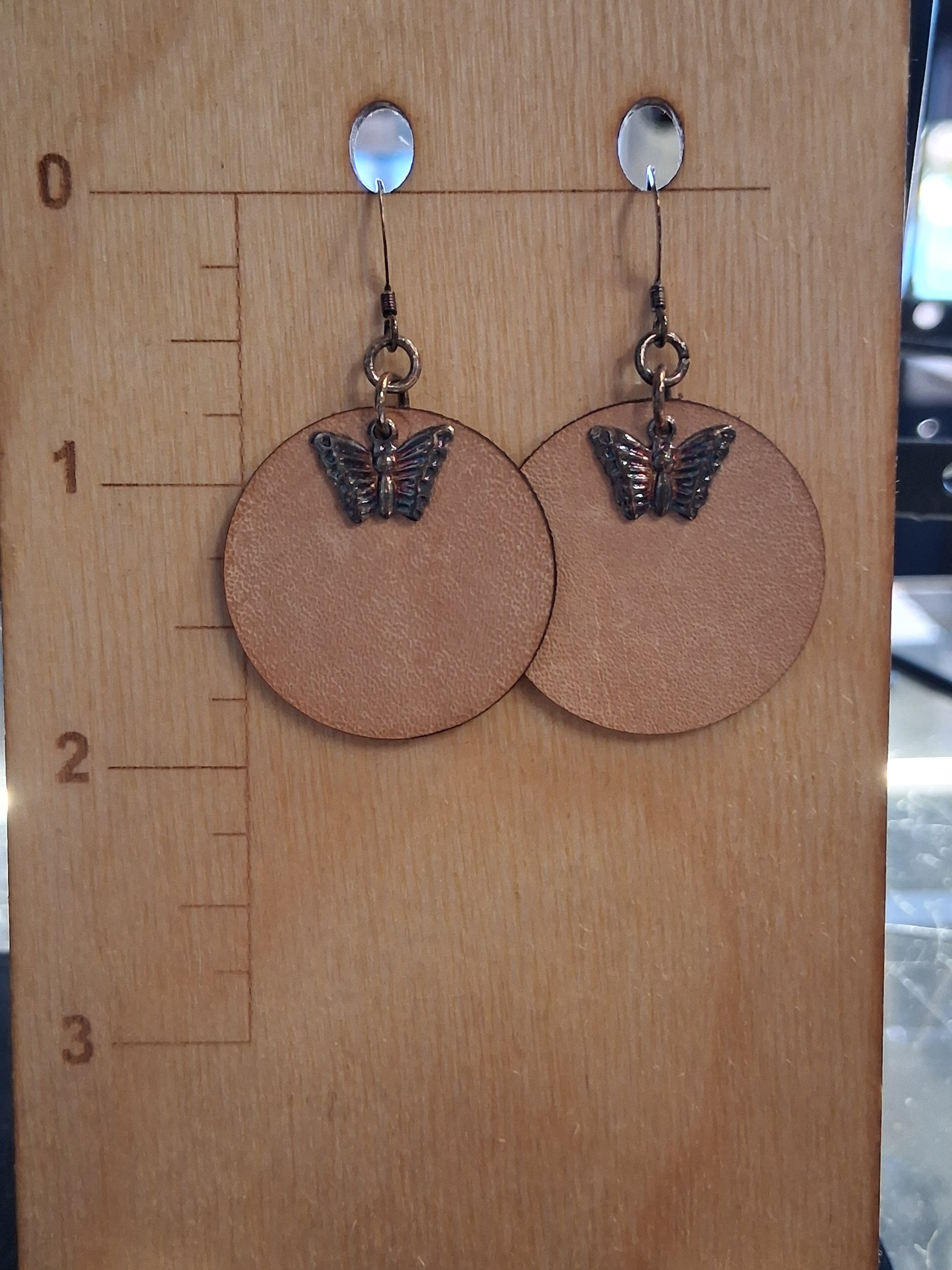 Leather earrings with butterfly charms on a wooden surface with a ruler for scale.