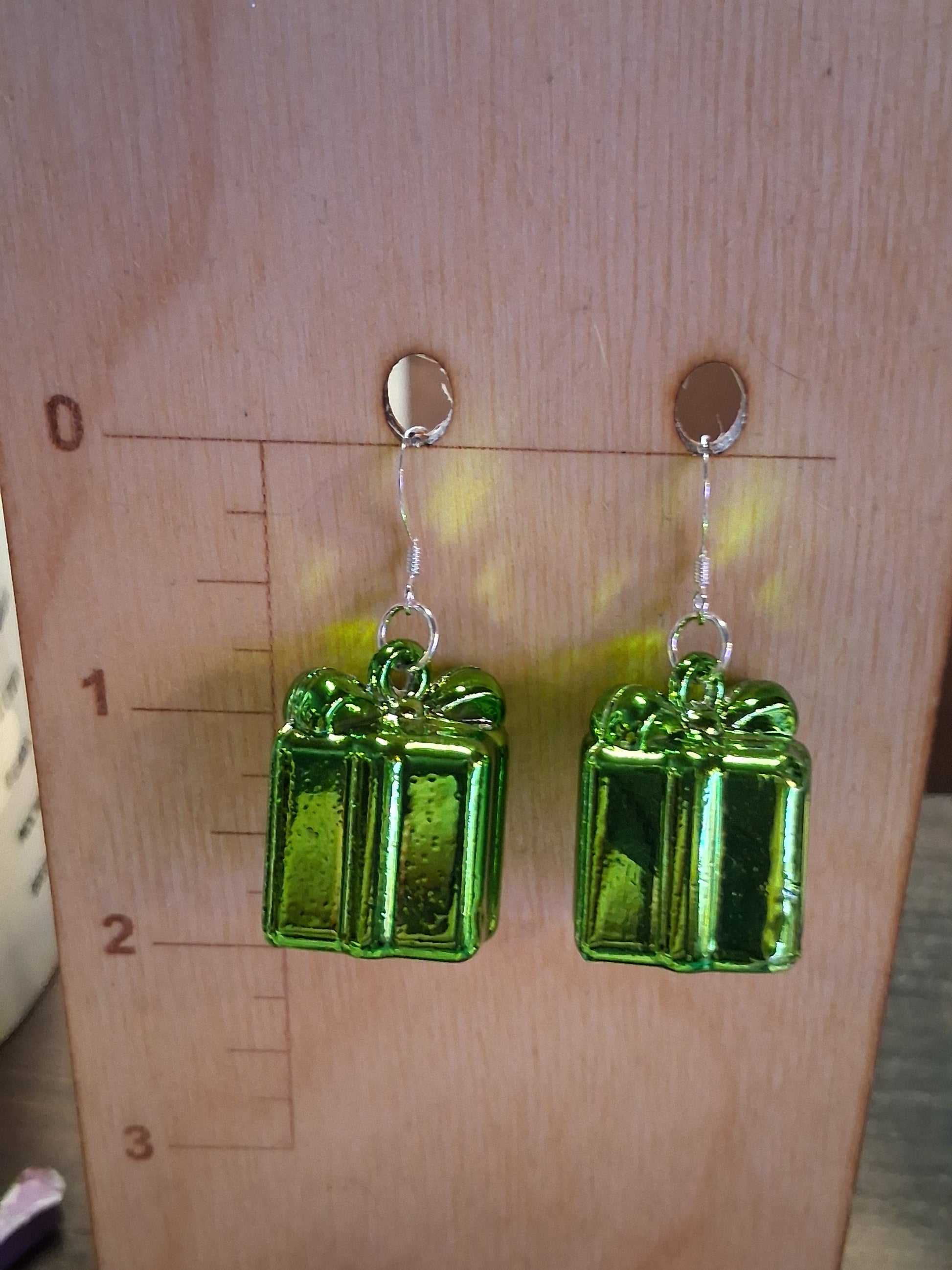 Green Christmas present charm earrings with metallic ribbon details and dark metal ear wires displayed on a wooden jewelry stand. Green rectangular present earrings on a wooden surface with a ruler for scale.