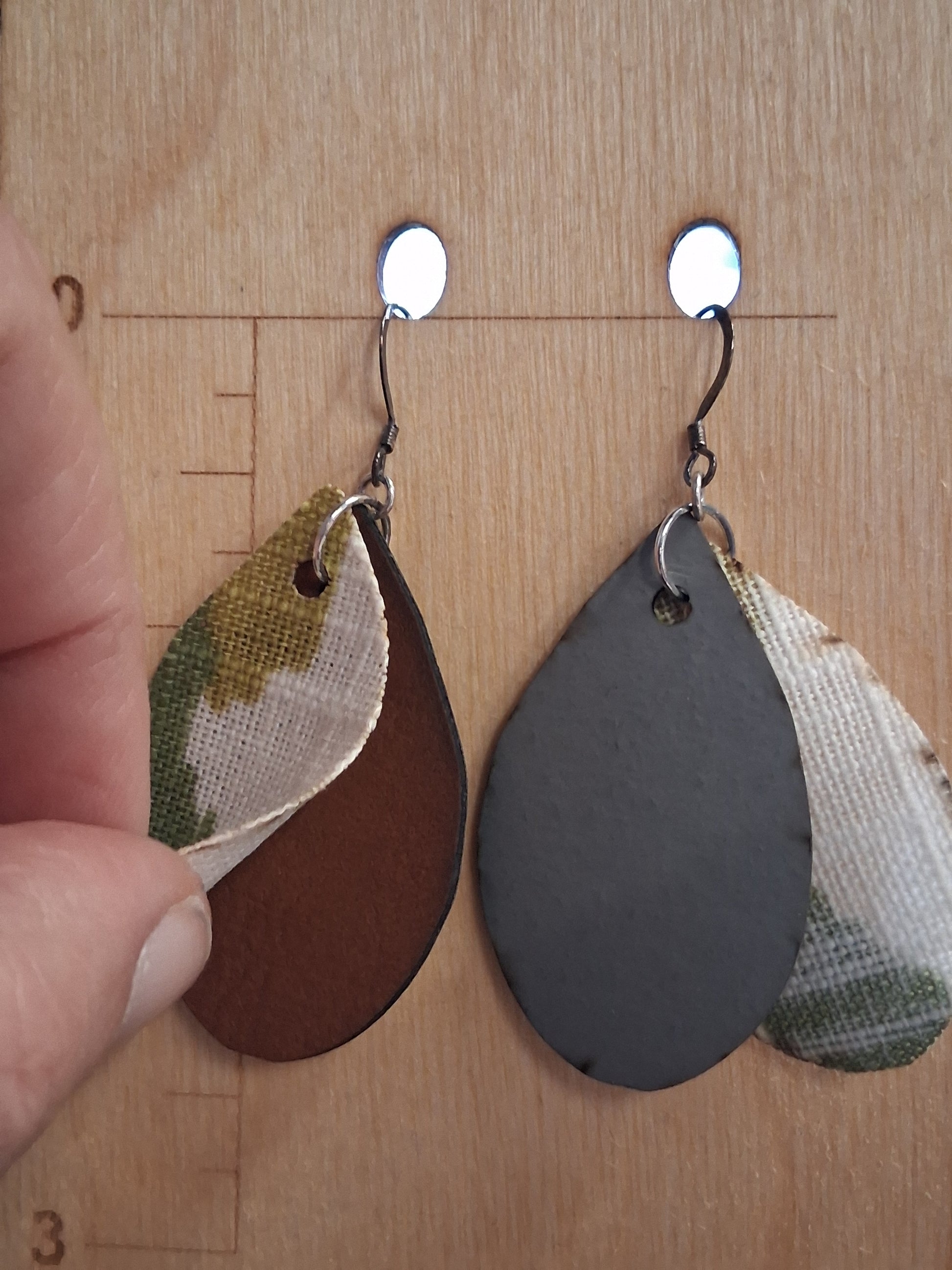 Pair of earrings with camouflage design fabric in front of a pleather back on a wooden surface with a ruler for scale.