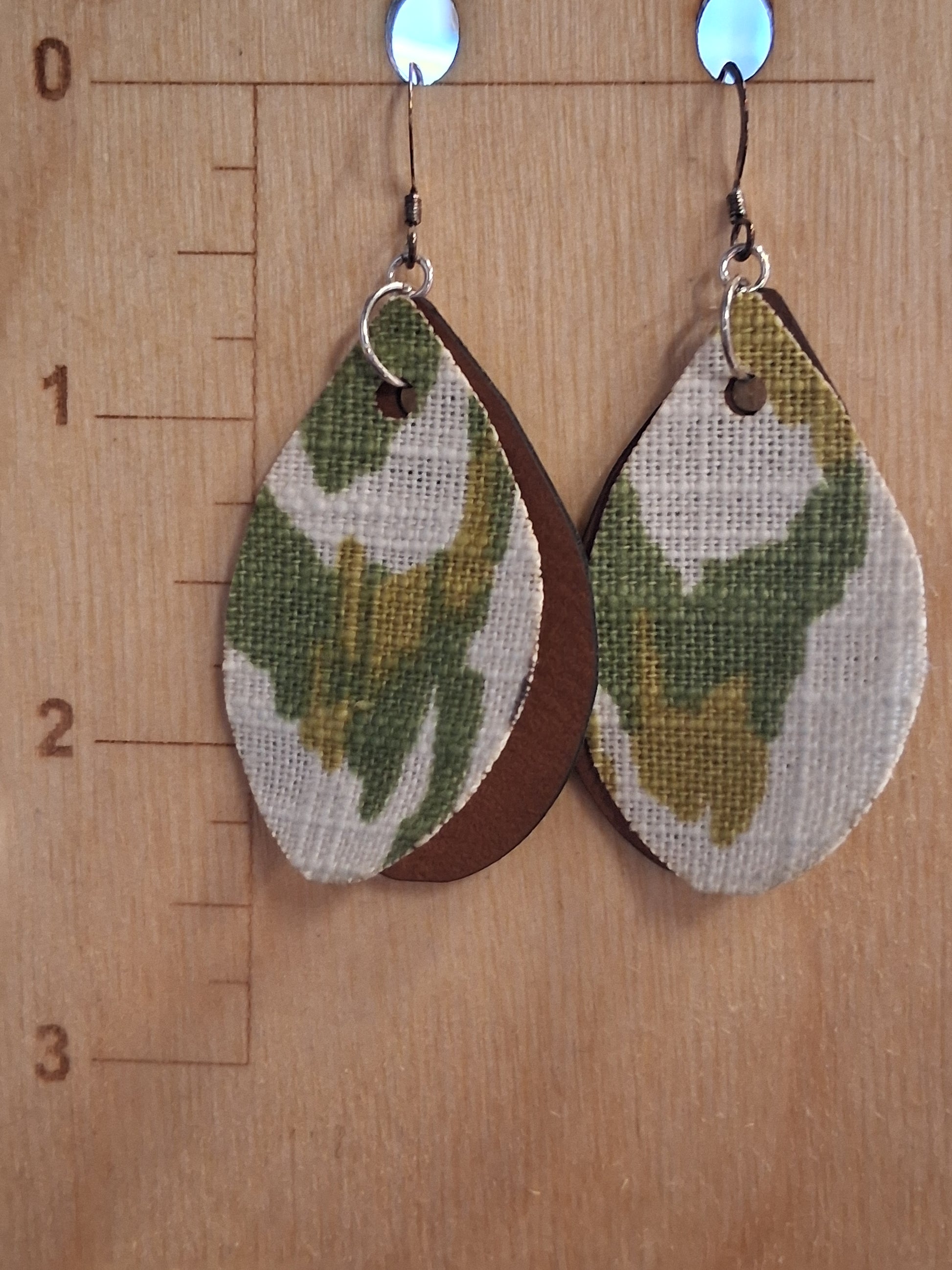 Pair of earrings with camouflage design fabric in front of a pleather back on a wooden surface with a ruler for scale.