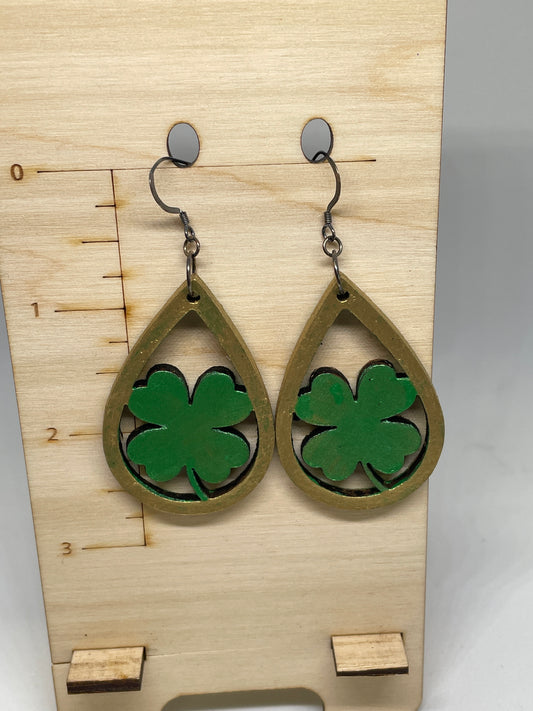 Handmade green shamrock leather earrings with antique-gold painted teardrop frames and nickel-free ear wires displayed on a wooden jewelry stand. Green four-leaf clover earrings on a wooden board with measurement markings.