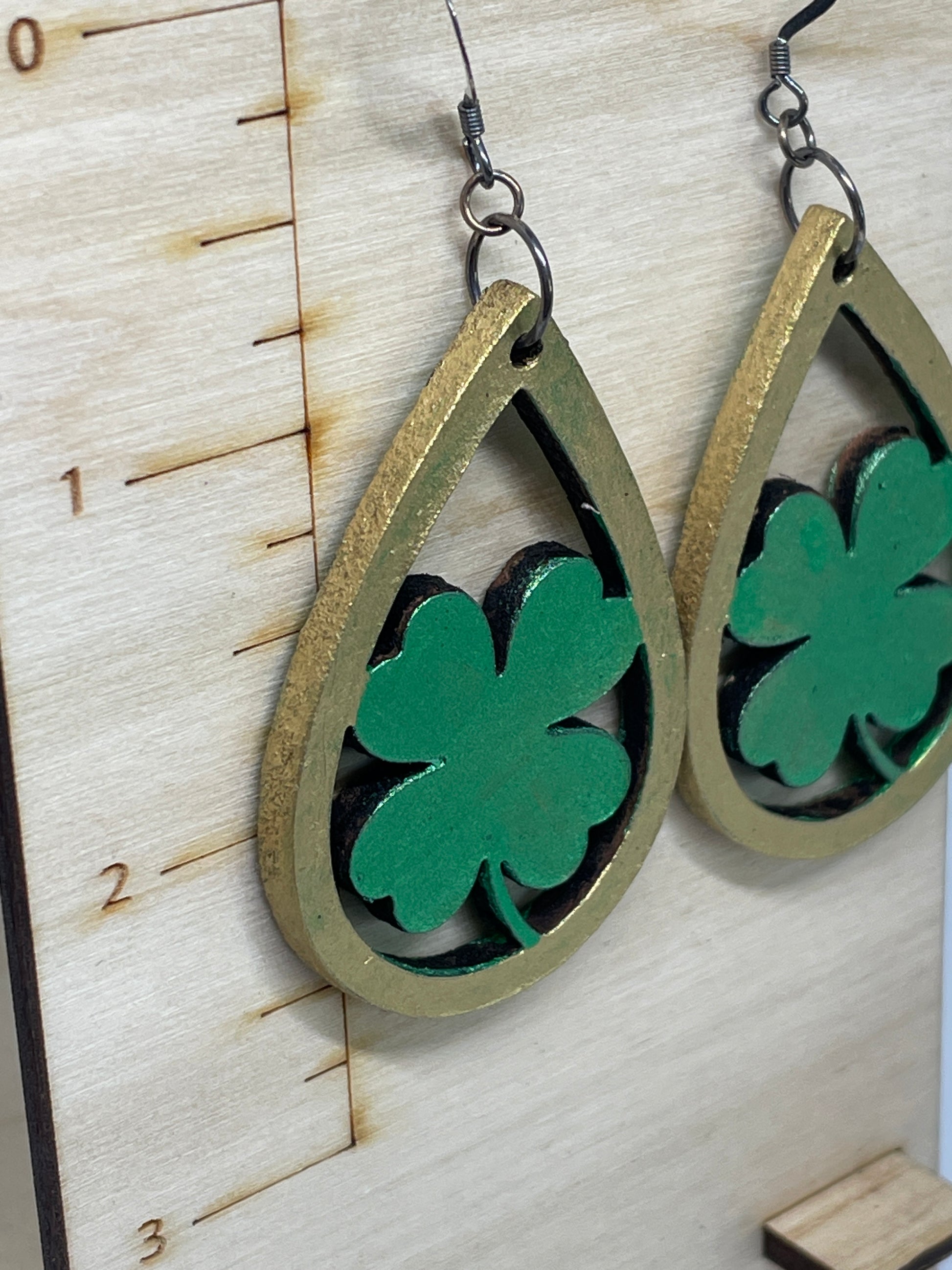 Close-up view of handmade shamrock leather earrings featuring green shamrock cutouts inside antique-gold painted teardrop frames. Pair of earrings with green clover leaves and gold teardrop shapes on a wooden surface.